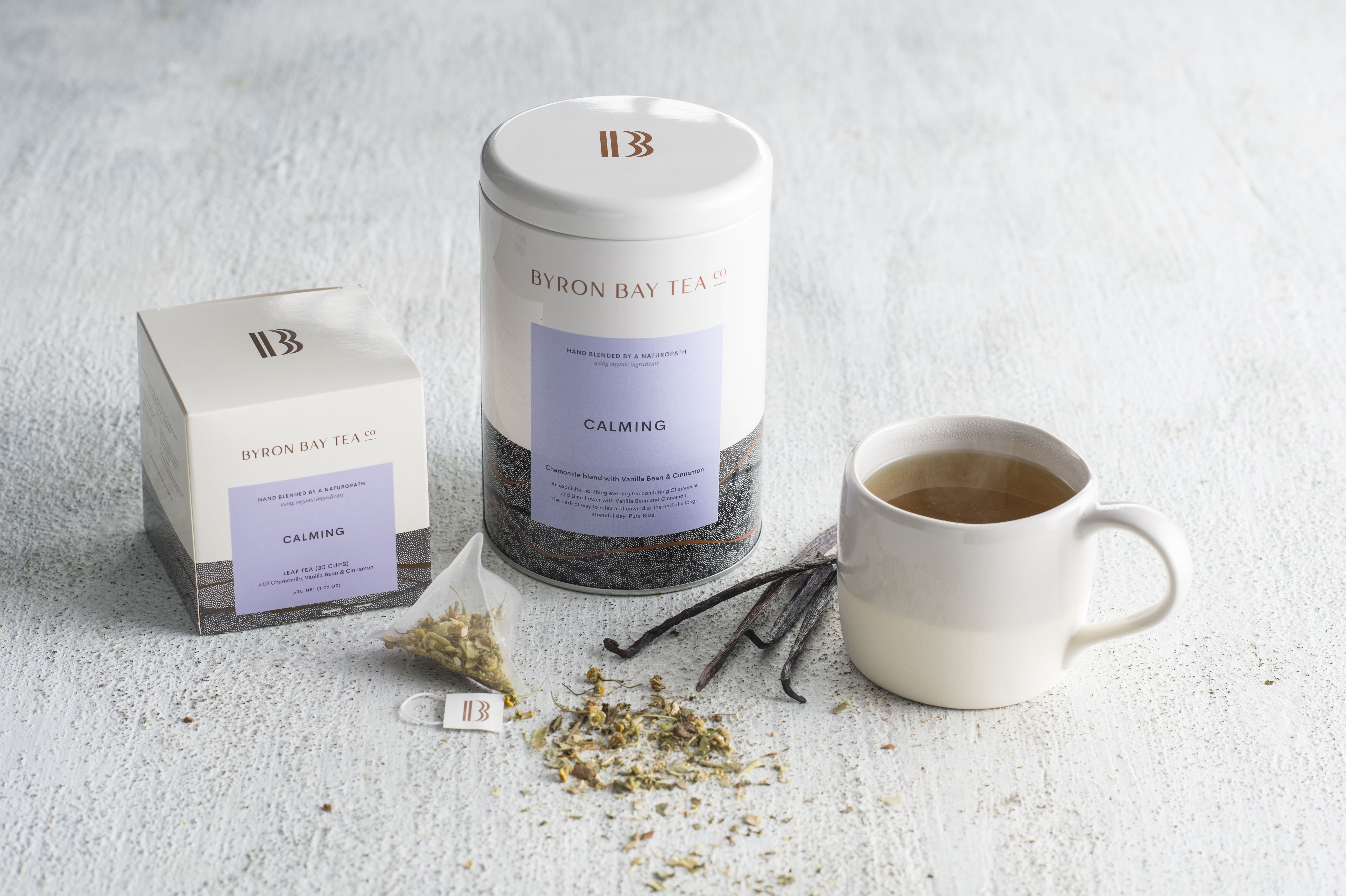 Box and tin of Byron Bay Tea Company calming tea with a teabag and tea leaf in the foreground and a brewed cup of tea in an organic grey cashmere mug next to it with vanilla sticks on a stone bench