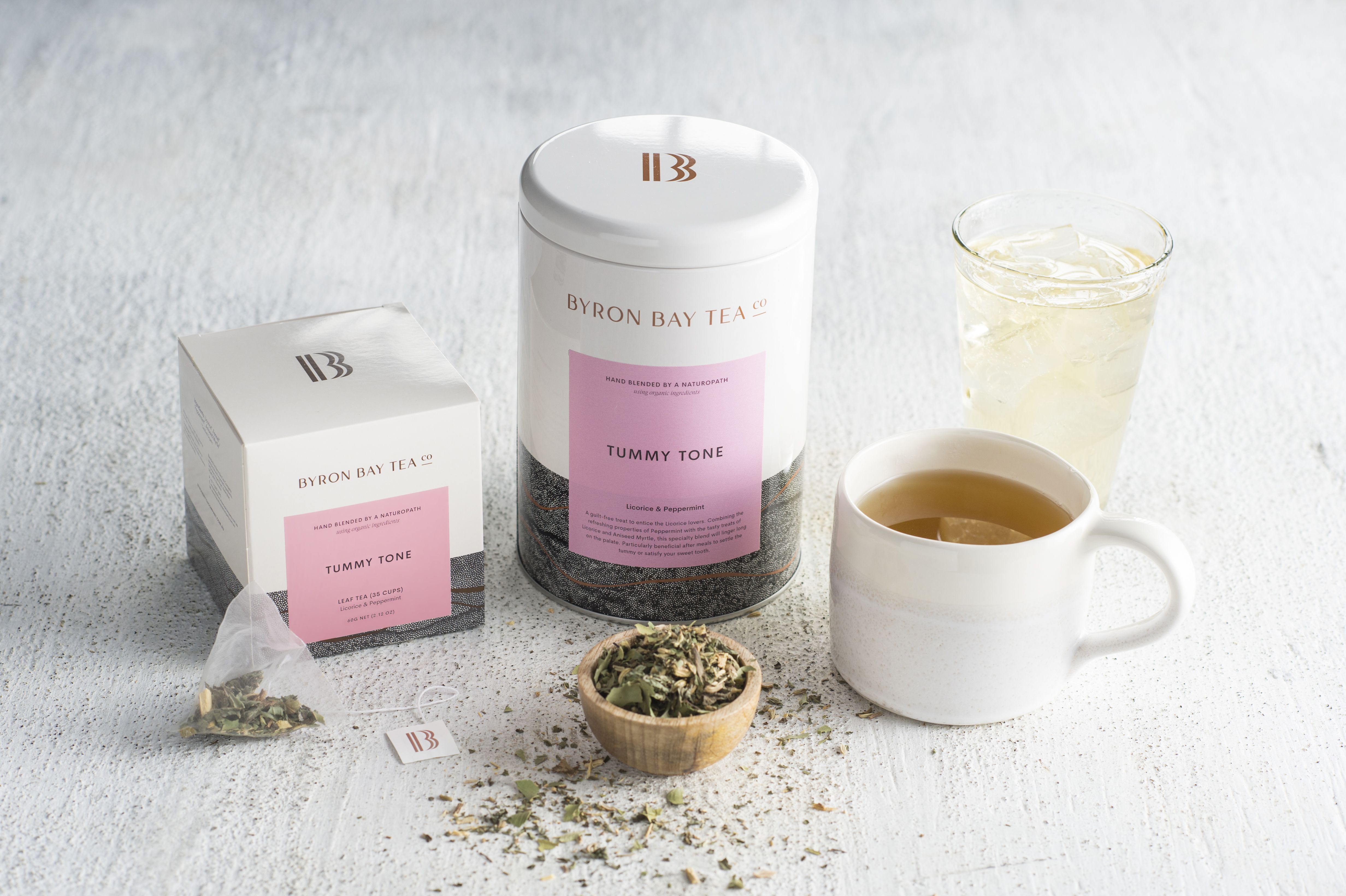 box and tin of byron bay tea company tummy tone tea with teabag and tea leaf in the foreground with organic cashmere mug with brewed tea and glass of. iced tea in the background on stone backdrop
