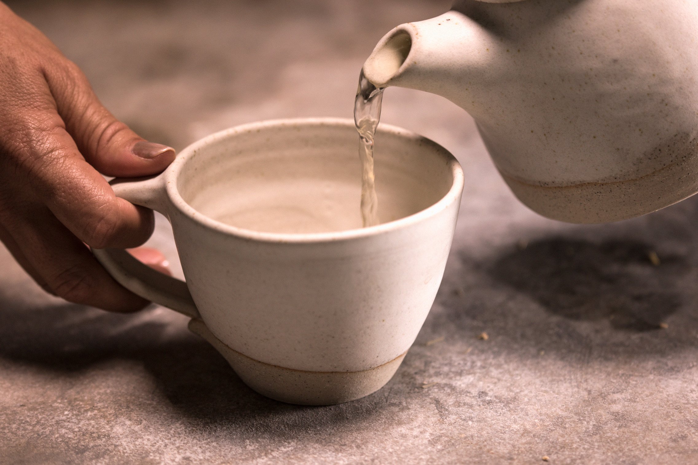 Pouring tea into a ceramic mug with matching ceramic teapot while holding on to mug on stone bench