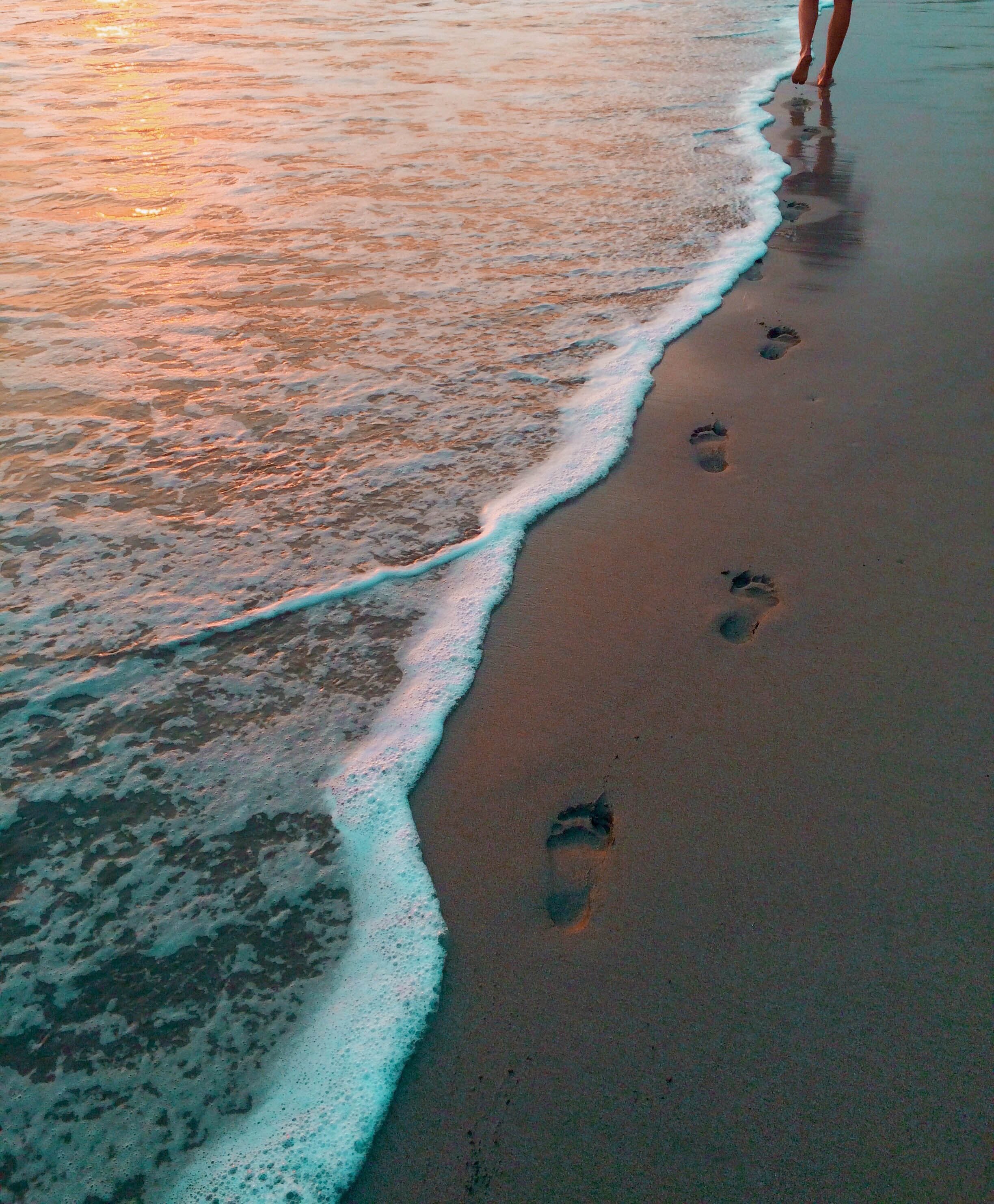 footprints being left behind person walking barefoot along the beach at sunset as a wave rises on the shore